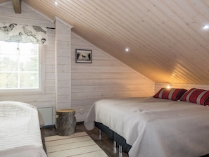 A cozy attic bedroom with two beds, a wooden ceiling, and a window with blinds.