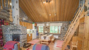 Interior acogedor de una cabaña de troncos con chimenea de piedra, una mesa de comedor de madera y una escalera.