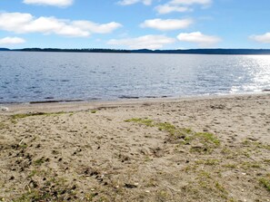 A sandy beach with a calm lake and a forested shoreline in the distance.