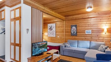 Ein gemütliches Blockhausschlafzimmer mit Holzbalkendecke, einem Fernseher auf einem Holztisch, einem grauen Sofa und einem kleinen Tisch mit einer Fernbedienung.