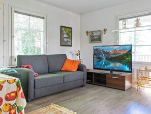 Una sala de estar con un sofá gris, un televisor de pantalla plana y un ventanal con vista a árboles.