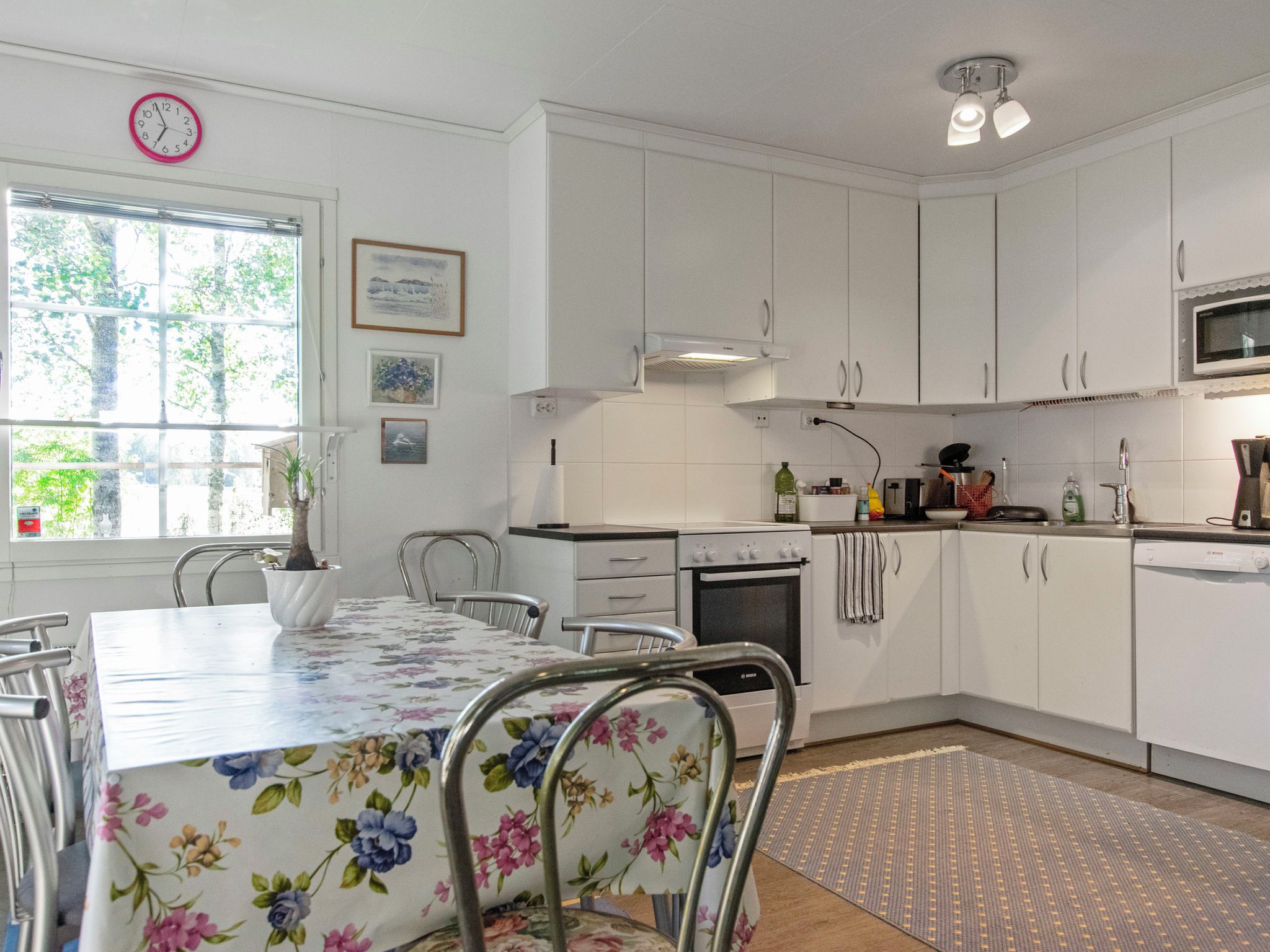 Eine Küche mit weißen Schränken, einem Esstisch mit Blumenmuster bedeckter Tischdecke und einer Uhr an der Wand.