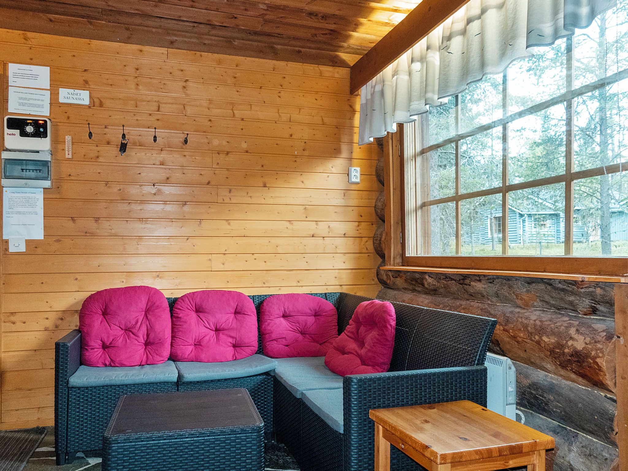 A wooden cabin interior with a sofa, cushions, a small table, and a window with a view of trees.