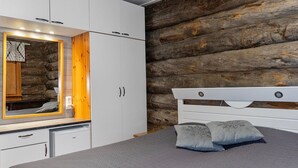 A bedroom with a bed, a wooden log wall, white cabinets, and a mirror with lights. - Raahenmaja by Interhome (Inari)