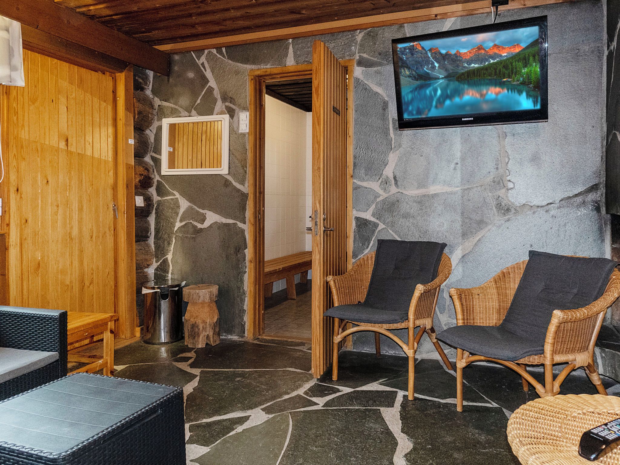 A stone-walled room with two wicker chairs, a television, and a wooden door leading to another room.