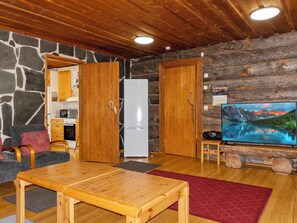 A cozy cabin interior with a stone wall, wooden ceiling, a dining table, a refrigerator, a television, and a kitchen area. - Raahenmaja by Interhome (Inari)