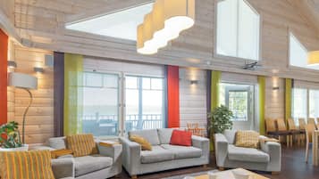 A spacious room with a high ceiling, wooden paneling, and large windows. There are three grey sofas with colorful cushions, a wooden coffee table, and a potted plant.