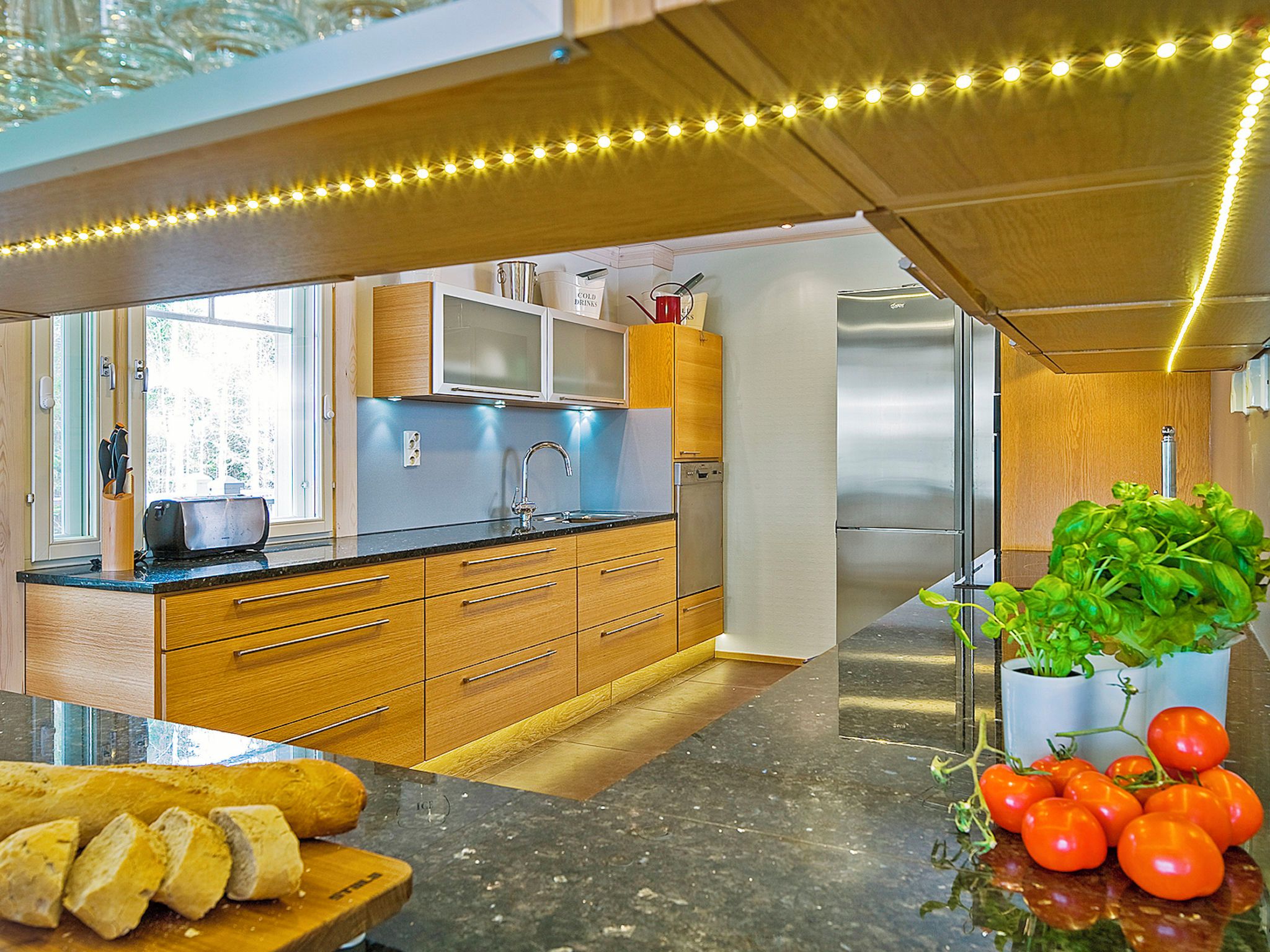 A modern kitchen with wooden cabinets, a stainless steel refrigerator, and a countertop with a plant and tomatoes.