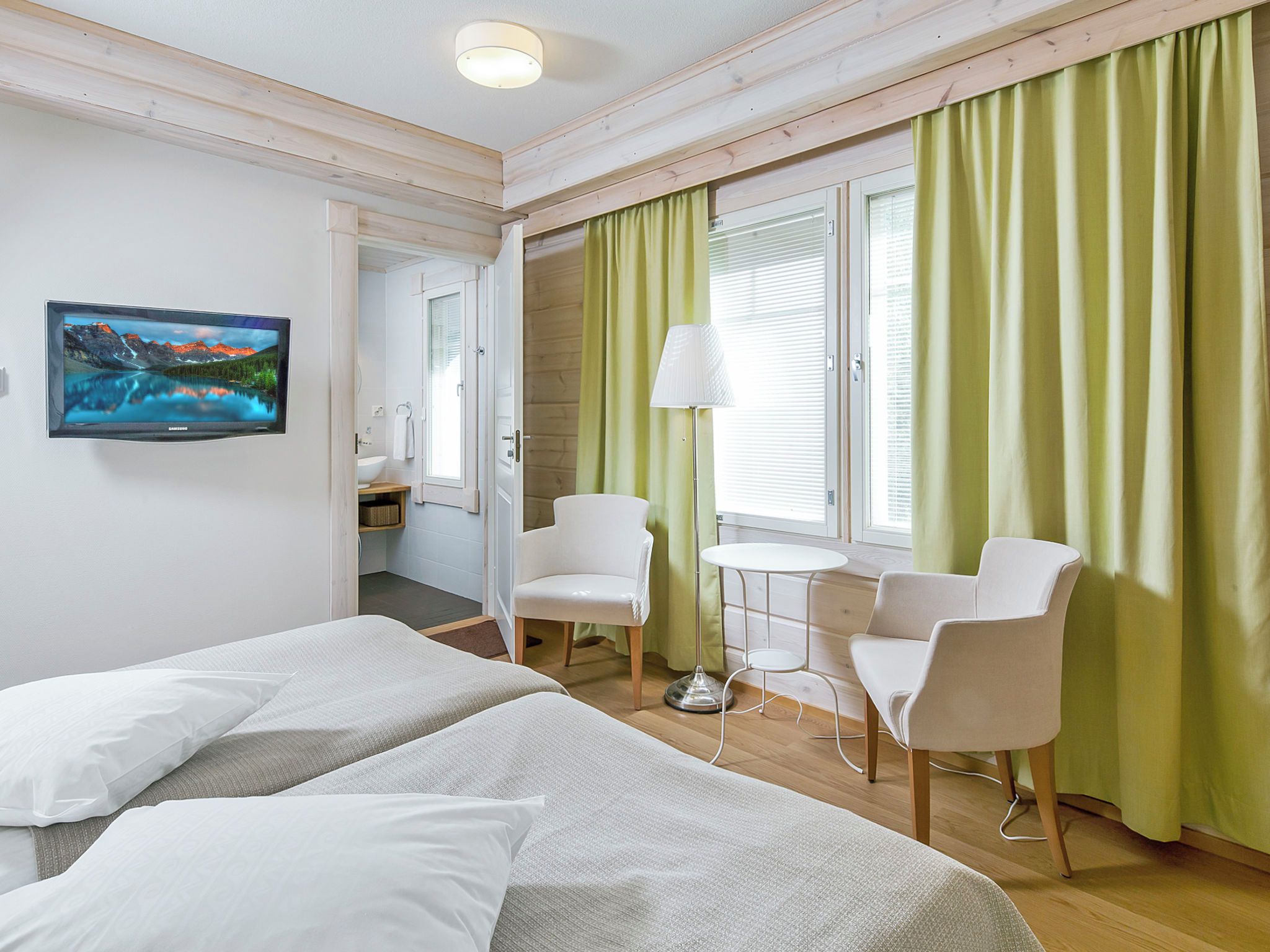 A hotel room with two beds, a small table, two chairs, a desk with a chair, a bathroom, and a painting on the wall.