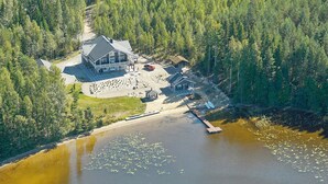An aerial view of a lodge surrounded by dense forest, a dock extending into a lake with floating vegetation, and a small boat docked nearby.