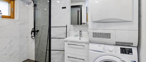 Casa de banho com chuveiro em box de vidro, pia com espelho, máquina de lavar roupa e sanita.