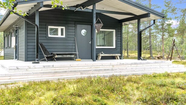 A dark wooden cabin with a covered porch, a bench, and a window overlooking a forested area.