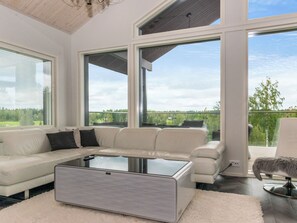 A modern living room with a sofa, a glass coffee table, and large windows offering a view of the outdoors.