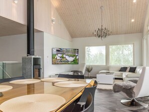 A modern interior with a dining area featuring a wooden table and woven placemats, a living room with a sofa and a television displaying ducks, and a kitchen with a built-in oven and a chimney. - Villa hill magia by Interhome (Kuhmoinen)