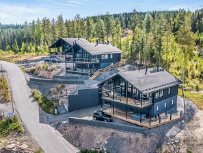 A modern multi-story house with a large balcony, surrounded by a forested landscape.