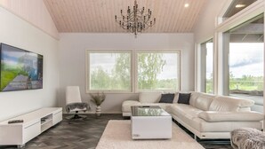 Un salon moderne avec un canapé blanc, une table basse et un téléviseur posé sur un meuble blanc. Il y a deux fenêtres équipées de stores, un lustre et un grand tableau accroché au mur.