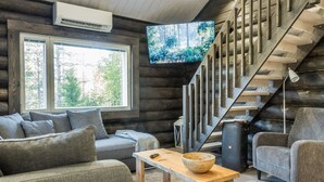 A log cabin interior with a sofa, a coffee table, a TV mounted on a wall, and a staircase.