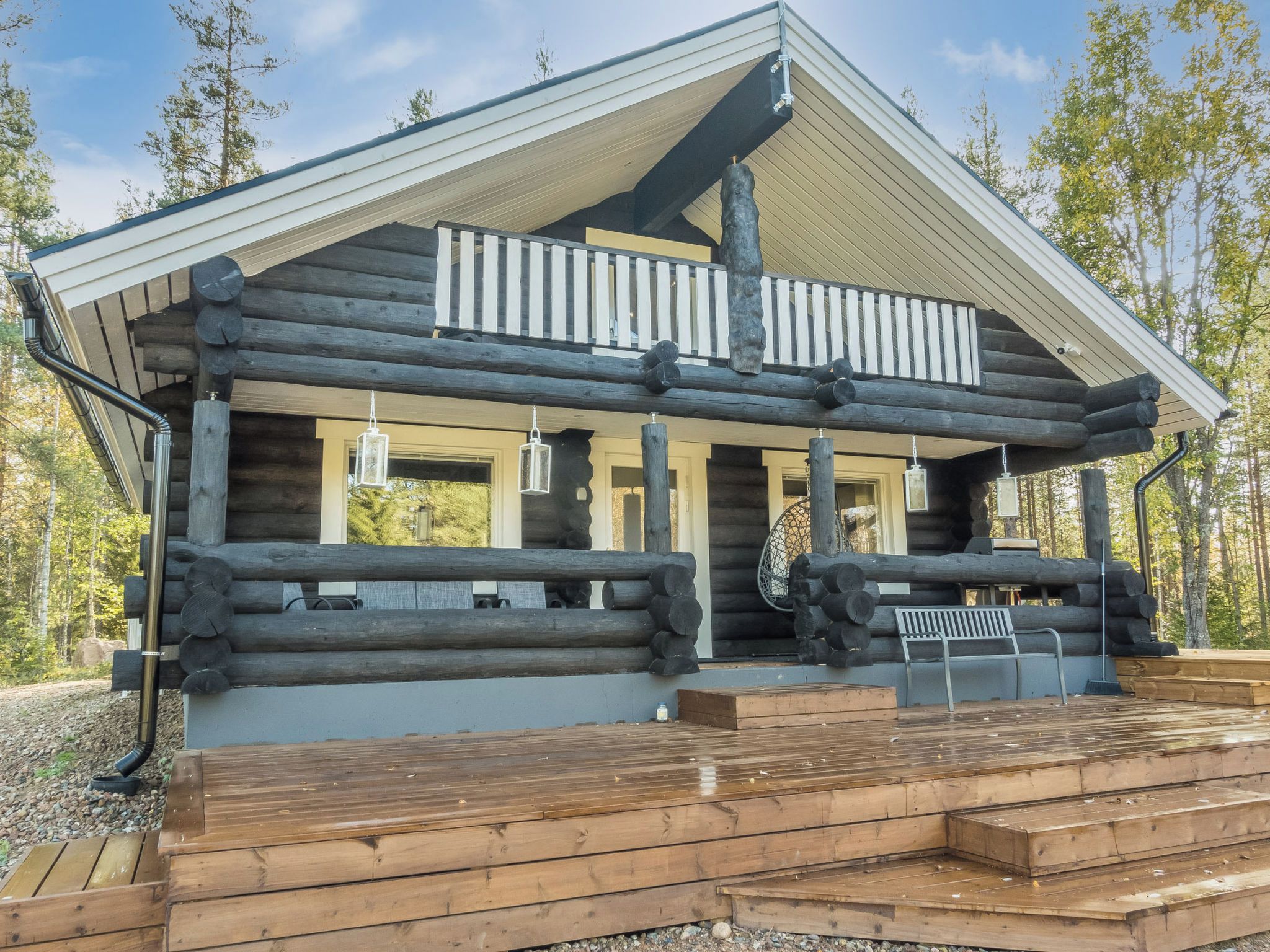 A log cabin with a balcony, wooden deck, and a view of the surrounding forest.