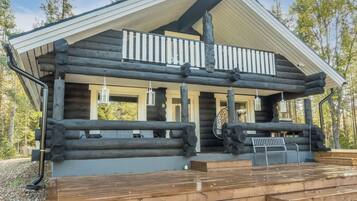 A log cabin with a balcony, wooden deck, and a view of the surrounding forest.