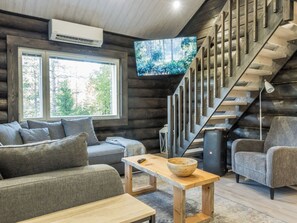 Interior de una cabaña de troncos con sofá, una mesa de madera, televisor montado en la pared y una escalera.