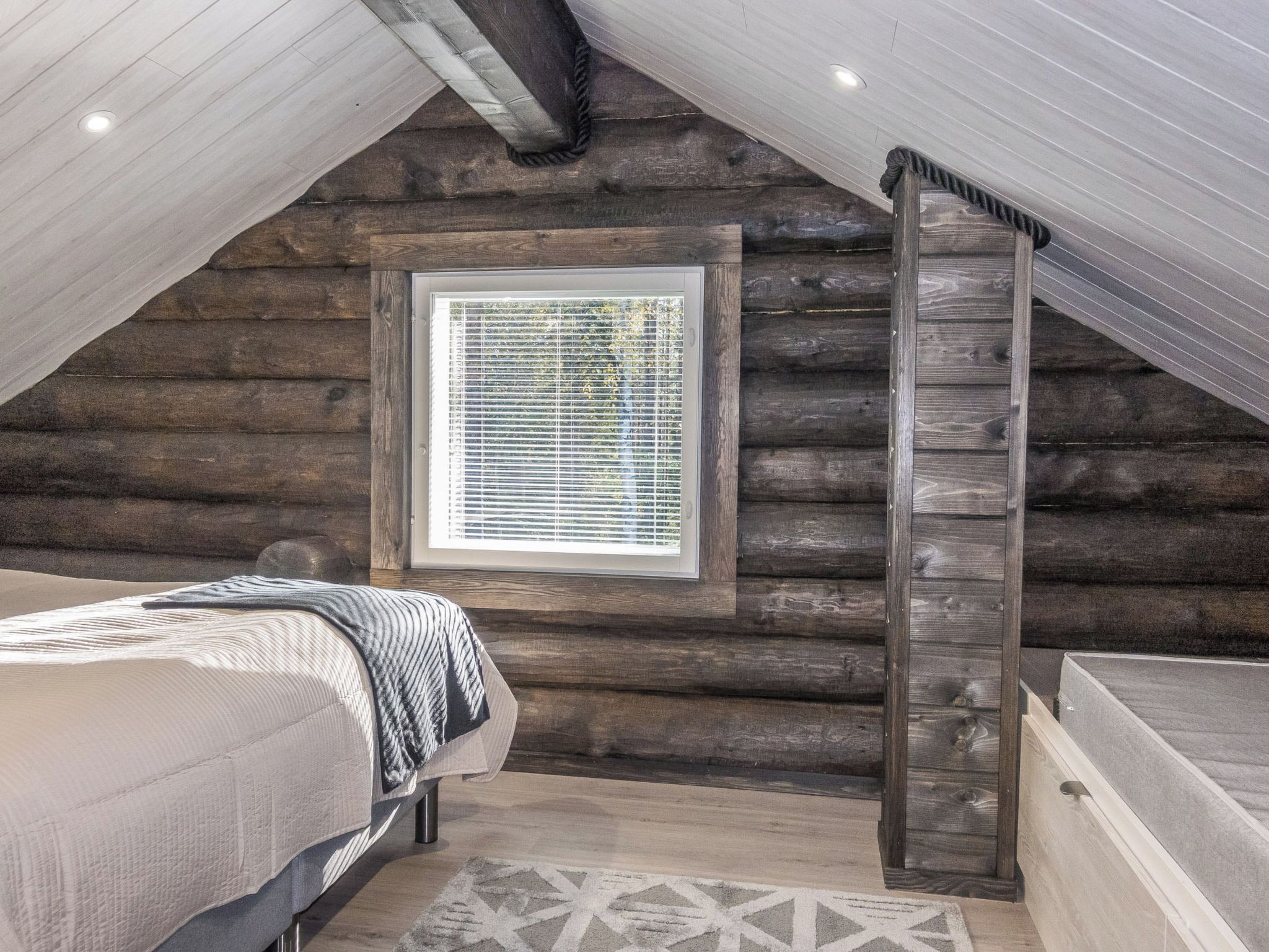 Ein Schlafzimmer in einer Blockhütte mit einem Bett, einem Fenster mit Jalousien, einer Bank und einer schrägen Decke.