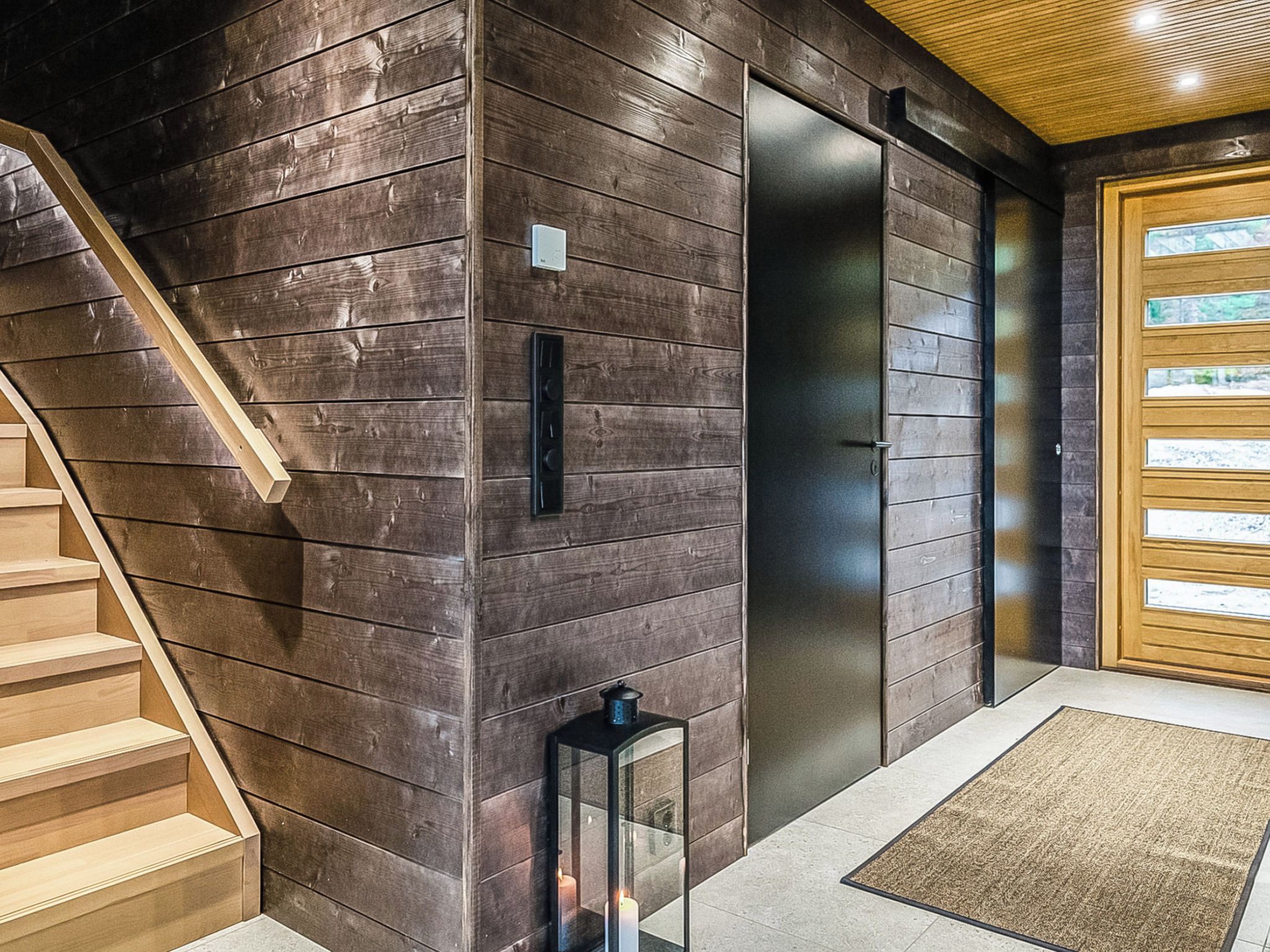 Un interior moderno con una escalera de madera, un aplique y una puerta con panel de vidrio.