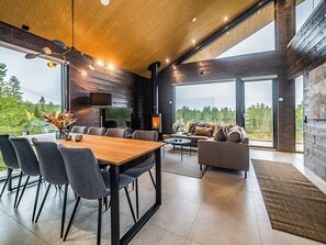 Et moderne interiør med spiseplads, siddepladser, en pejs og store vinduer med udsigt over naturen.