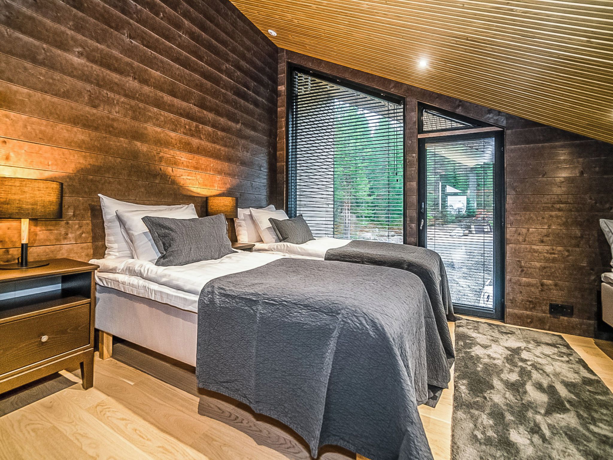 Une chambre avec un mur en lambris de bois, un lit avec de nombreux oreillers, une table de chevet et une grande fenêtre avec des stores.