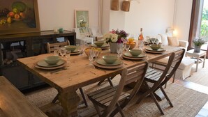Een eetruimte met een houten tafel voor zes personen, omringd door stoelen. Aan de muur hangt een schilderij, er is een hanglamp en een raam met jaloezieën.
