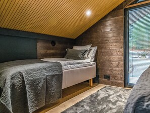 A bedroom with a sloped ceiling, a bed with grey bedding, a wooden floor, and a large window with blinds.