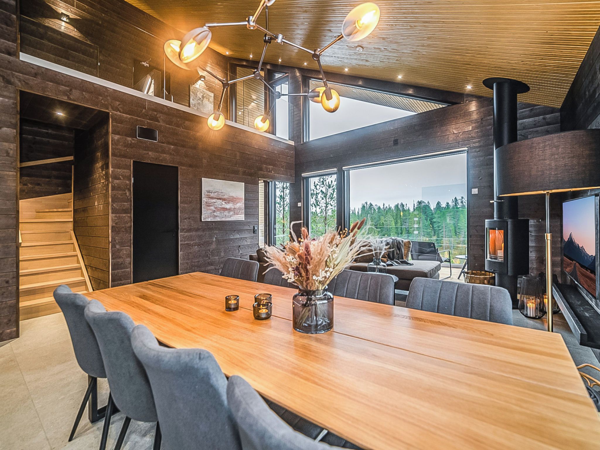 Um interior moderno com uma mesa de jantar de madeira, cadeiras estofadas cinzentas, lareira e uma grande janela com vista para árvores.