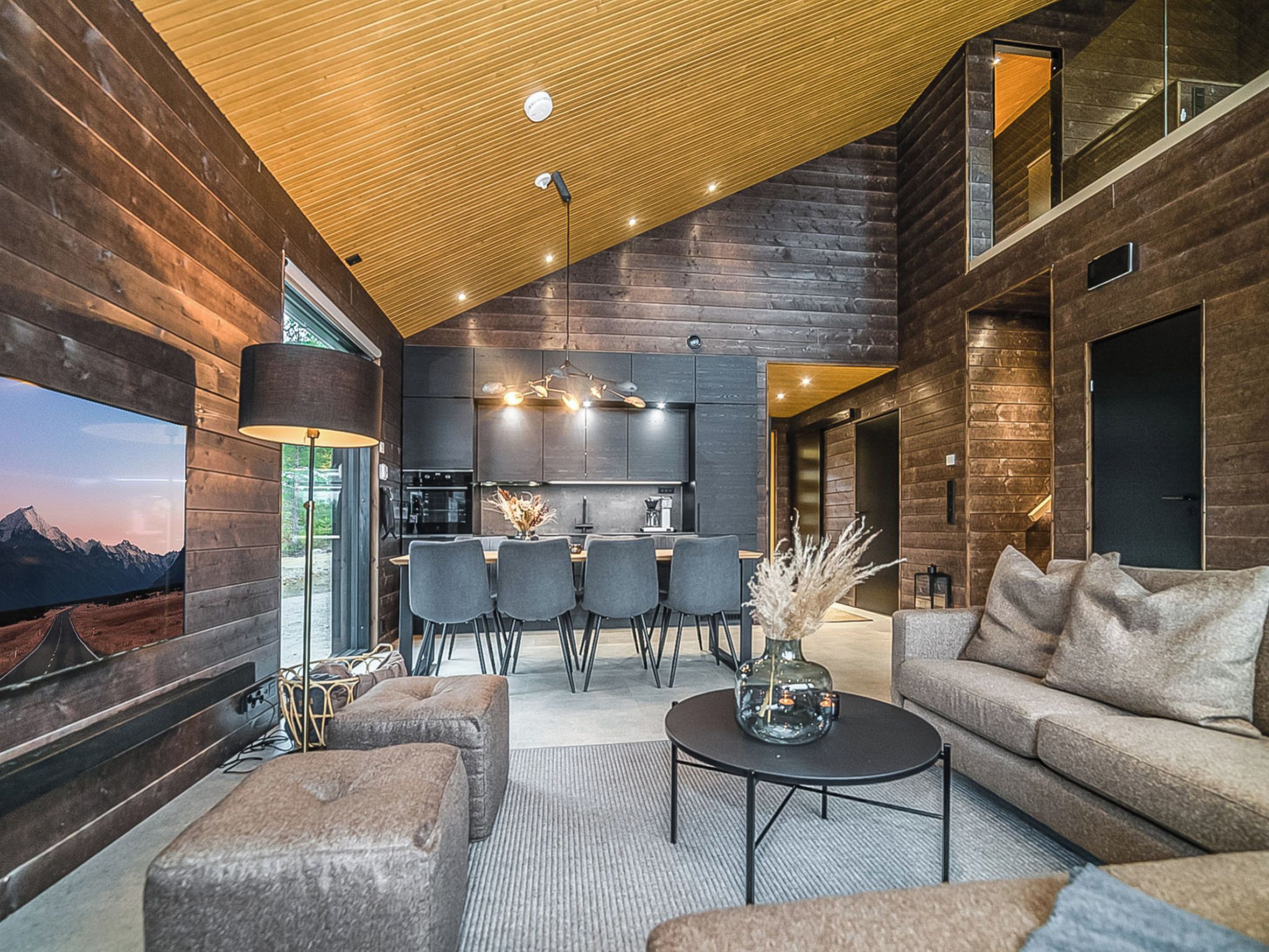 Un intérieur moderne avec un plafond en bois, un coin cuisine, un espace salon avec un canapé et des fauteuils, et un grand téléviseur à écran plat fixé au mur.