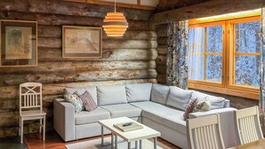 Ein gemütliches Blockhaus-Interieur mit grauem Sofa, weißen Stühlen, einem kleinen Tisch und einem Fenster mit Blick auf Bäume.