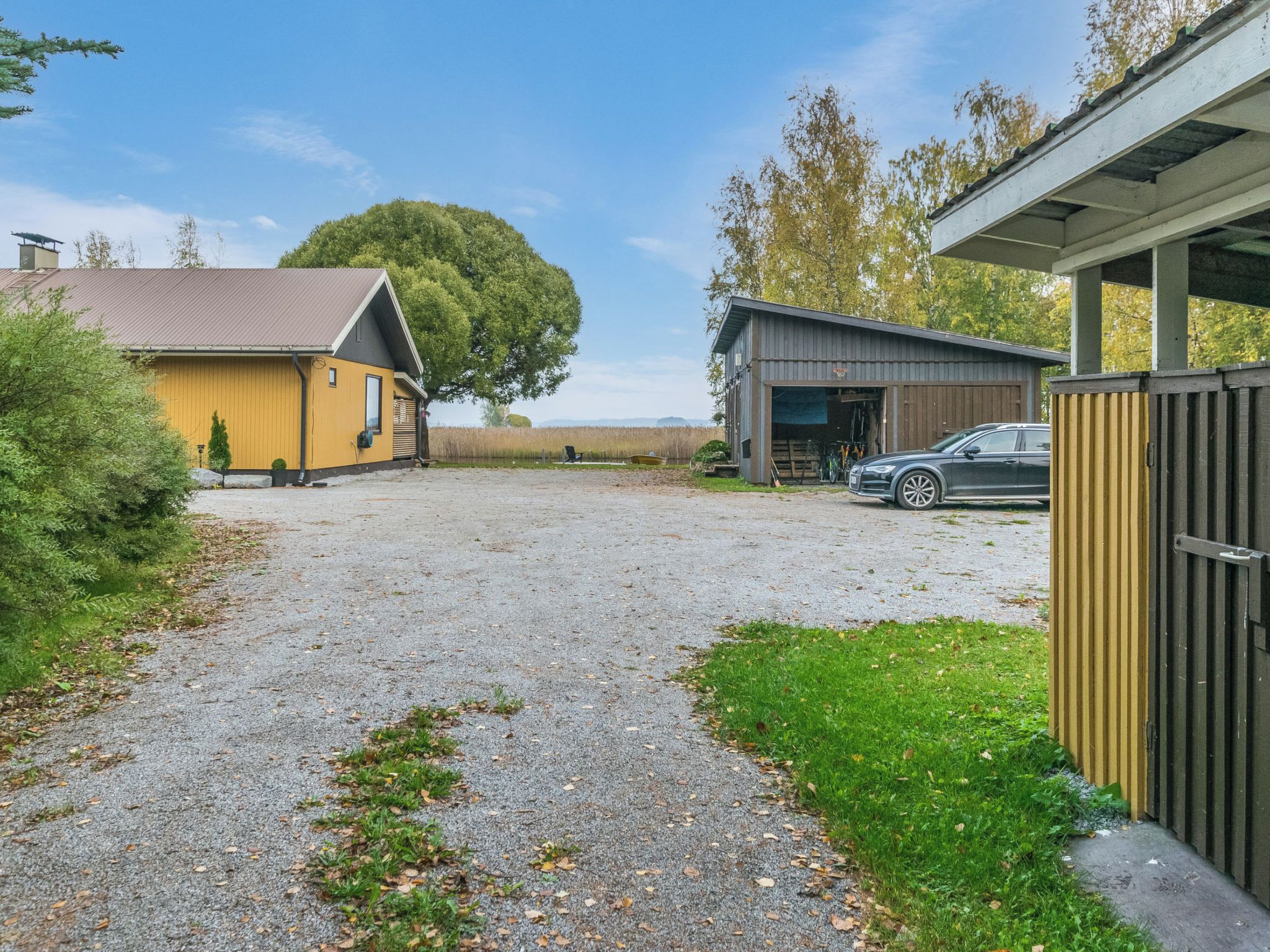 A gravel driveway leading to a yellow building with a metal roof, a black car parked in front, and a large tree in the background.