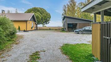 A gravel driveway leading to a yellow building with a metal roof, a black car parked in front, and a large tree in the background.