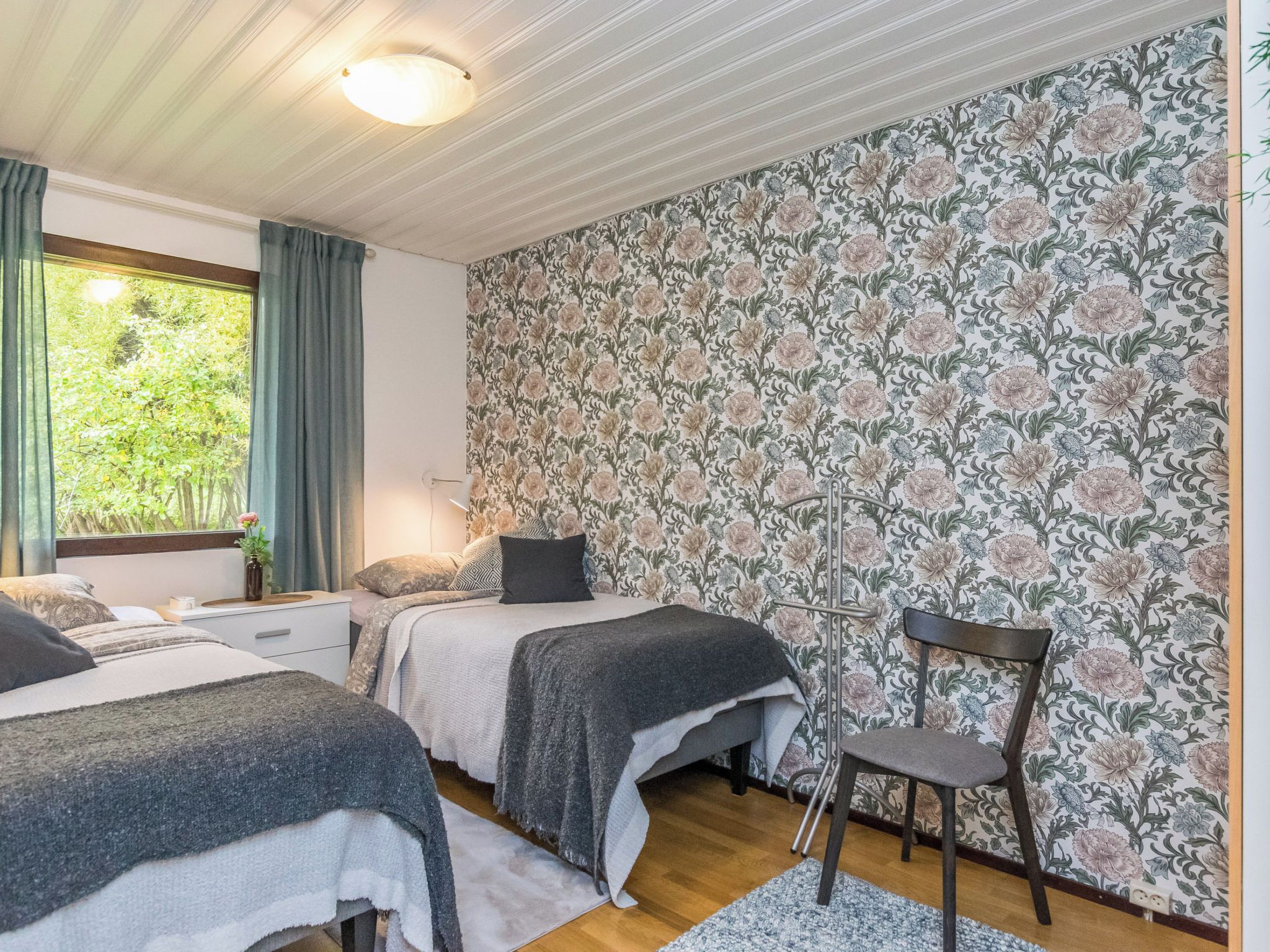 Ein Schlafzimmer mit zwei Betten, einem Sessel, einem Fenster mit Vorhängen und floralem Wandpapier.