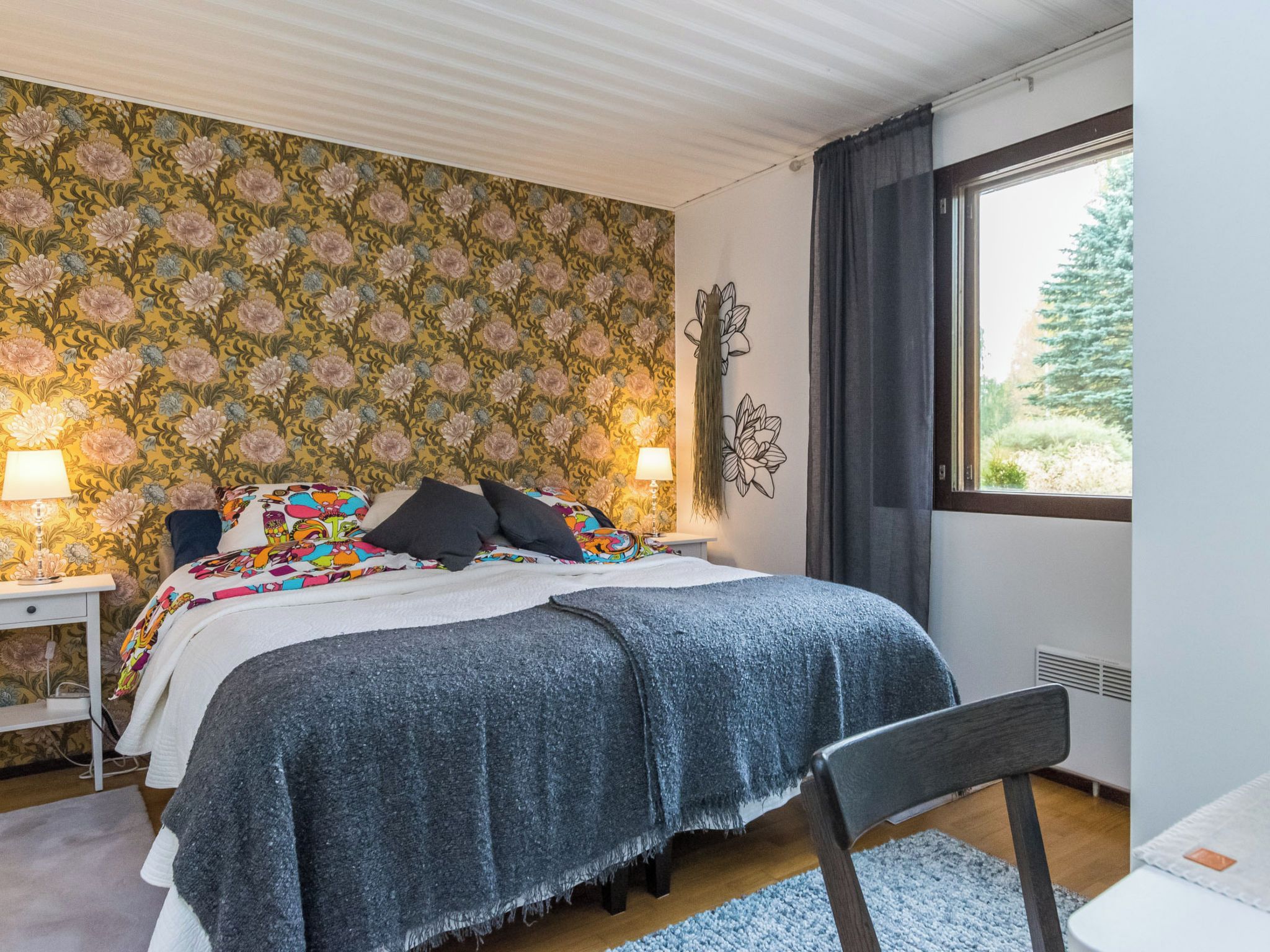 A bedroom with a bed, bedside tables, lamps, a window with curtains, and a wall with floral wallpaper.