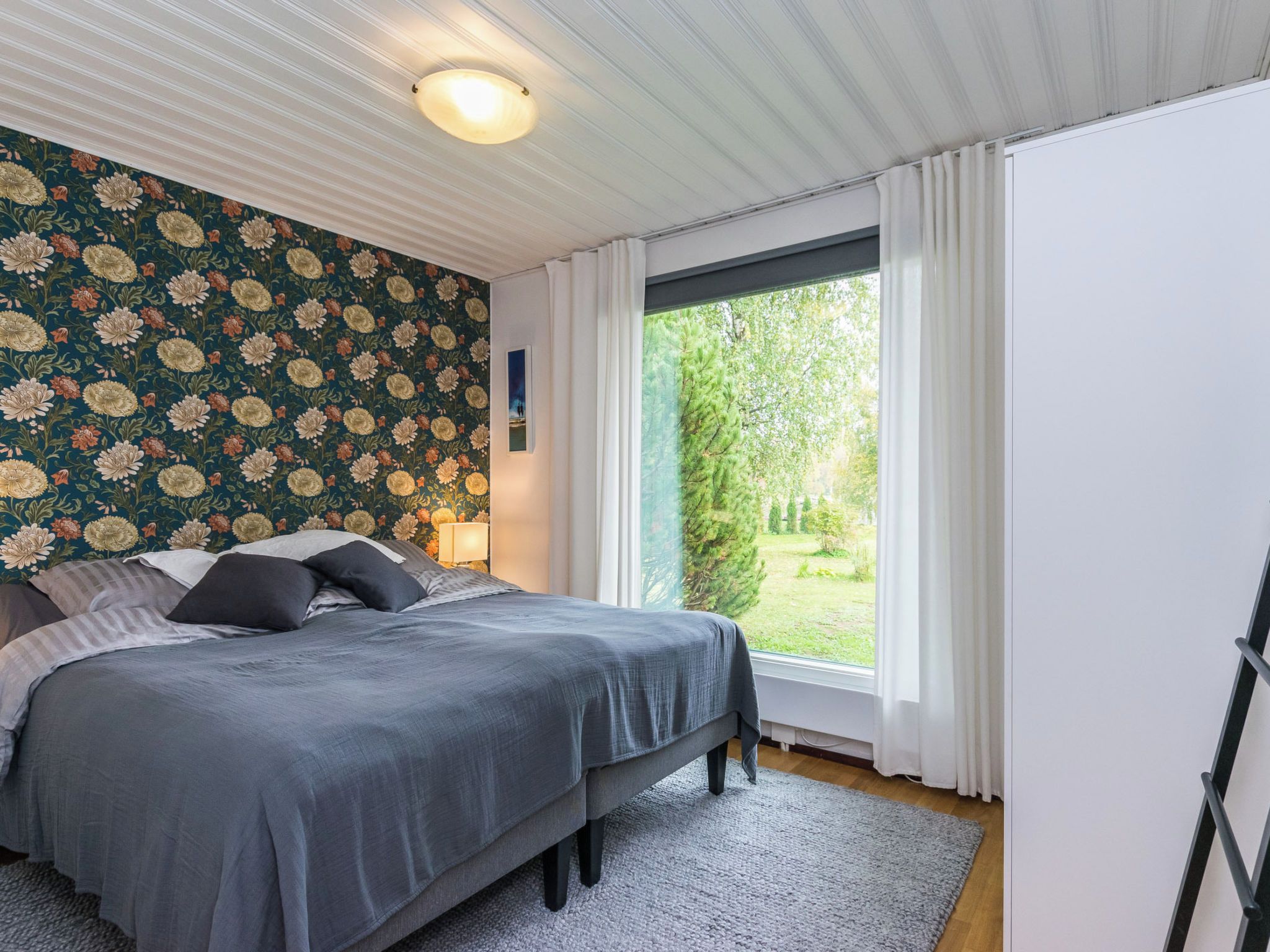 Une chambre à coucher avec un lit, une table de chevet, une lampe, une fenêtre avec des rideaux et une vue sur la verdure à l’extérieur.