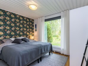 A bedroom with a bed, a nightstand, a lamp, a window with curtains, and a view of greenery outside.