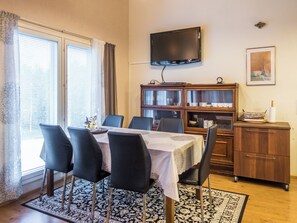 Een eetruimte met een tafel voor vier personen, stoelen, een aan de muur gemonteerde televisie, een kast met glazen deuren en een raam met gordijnen.