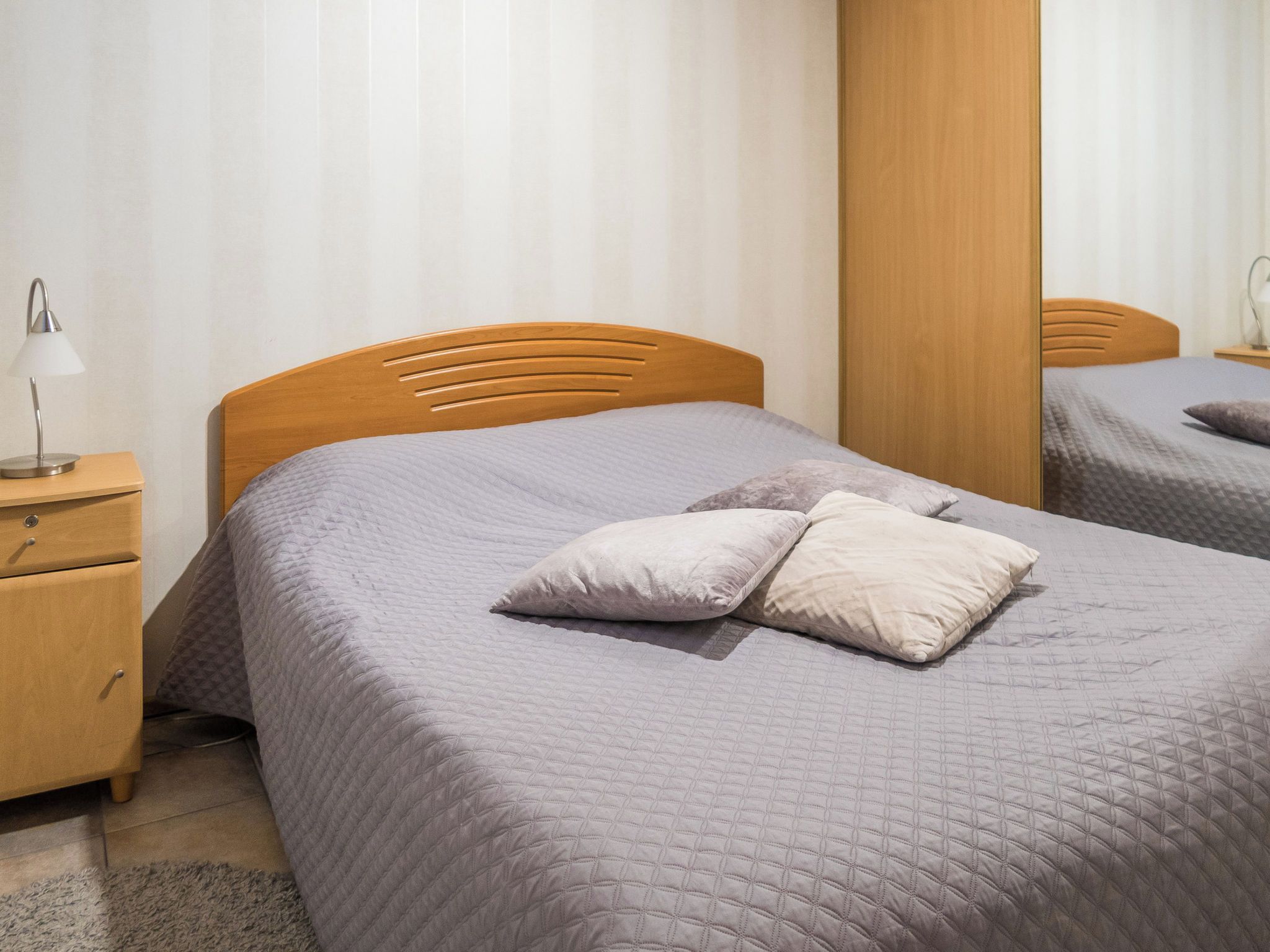 Una cama bien hecha con cabecera de madera, una mesita de noche con lámpara y un espejo en la pared.