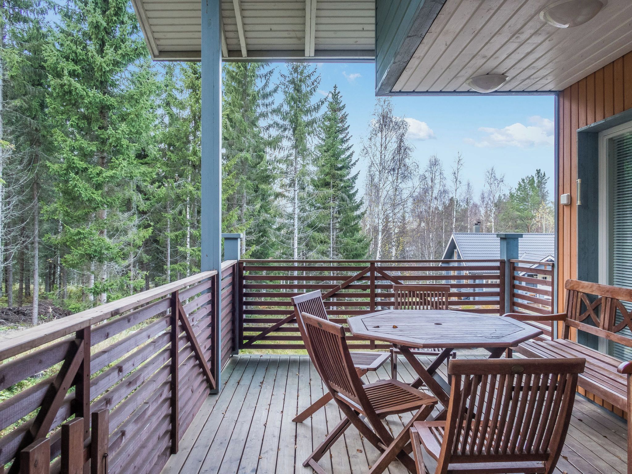 A wooden deck with a table and chairs, surrounded by a railing, overlooking a forested area.