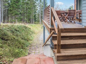 Une terrasse en bois avec des meubles d’extérieur, un escalier menant à une zone abritée et un paysage boisé en arrière-plan.