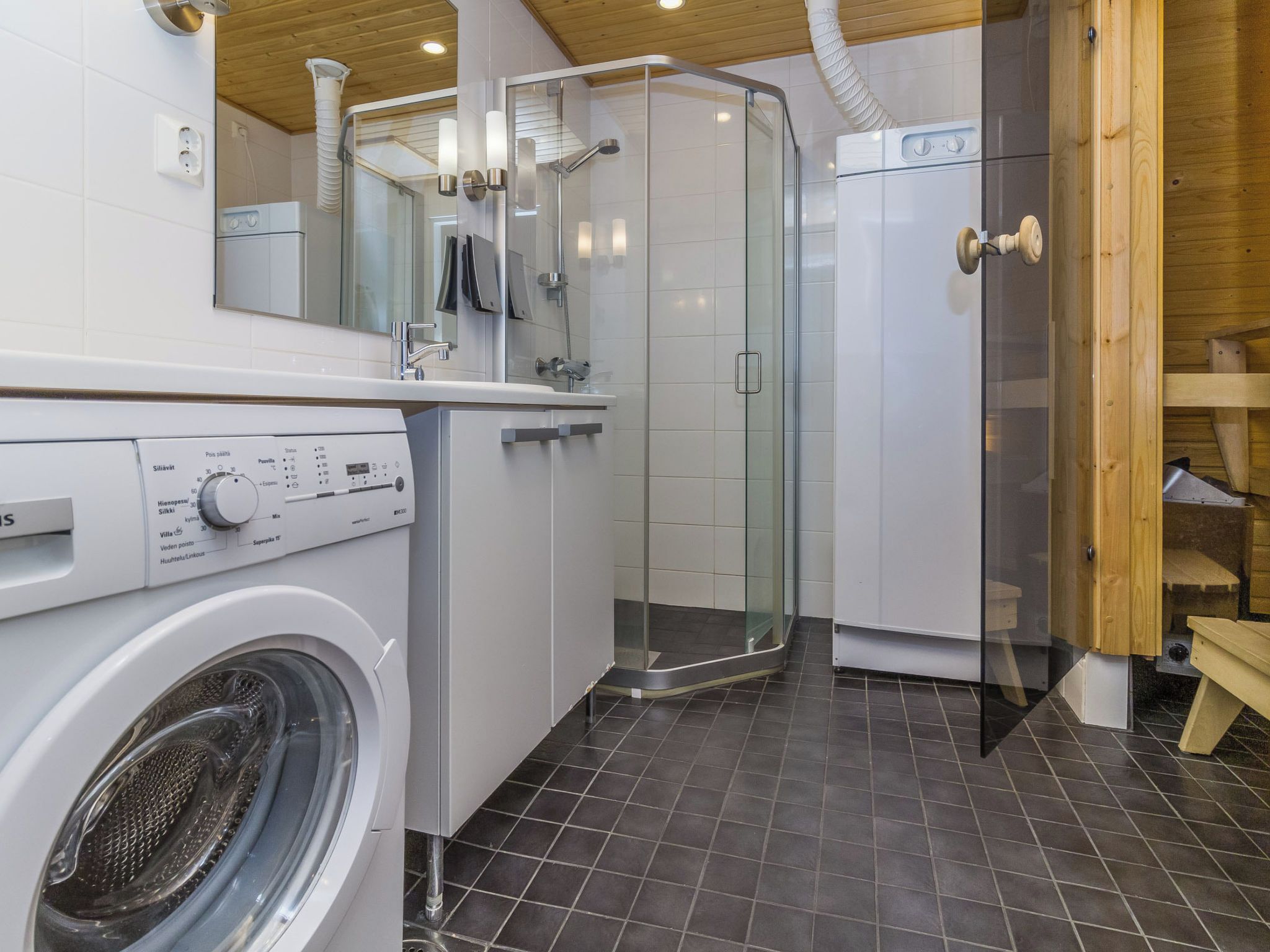 A compact bathroom with a washing machine, a glass shower enclosure, and a wooden bench.