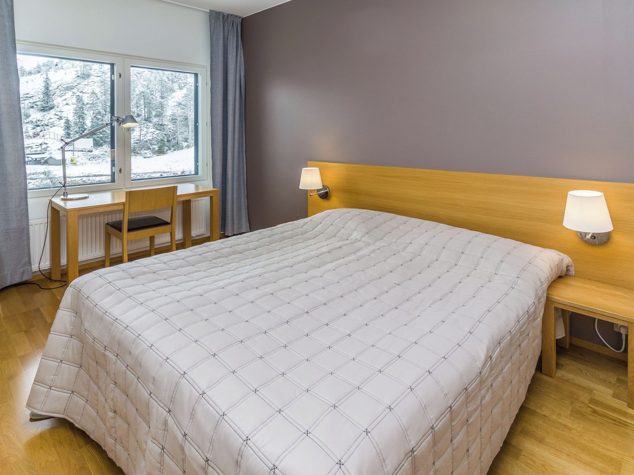 Une chambre d’hôtel avec un grand lit, un bureau, deux lampes de chevet et une fenêtre donnant sur des arbres couverts de neige.
