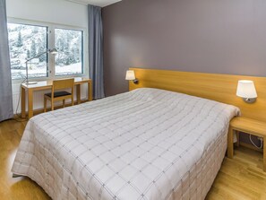 Une chambre d’hôtel avec un grand lit, un bureau, deux lampes de chevet et une fenêtre donnant sur des arbres couverts de neige.