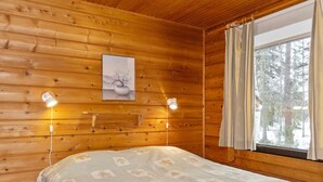 A wooden cabin room with a bed, two bedside lamps, a framed picture on the wall, and a window with curtains.