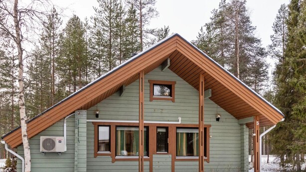 A green wooden cabin with a pitched roof, large windows, and a small porch area.
