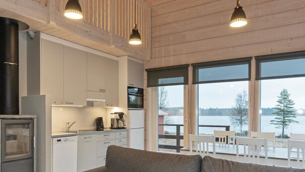 A modern kitchen with white cabinets, a built-in oven, and a coffee maker. There is a gray sofa and a dining table with chairs in the living area. Large windows offer a view of a lake and trees.