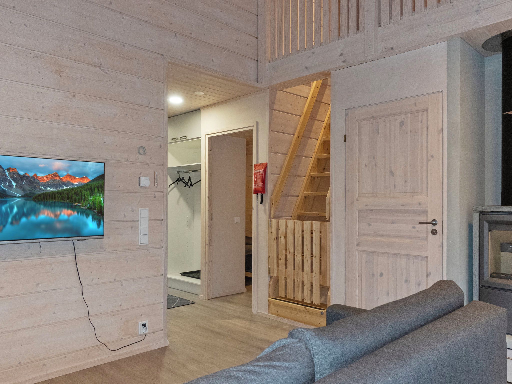A wooden interior with a television mounted on the wall, a grey sofa, and a staircase leading to an upper level.